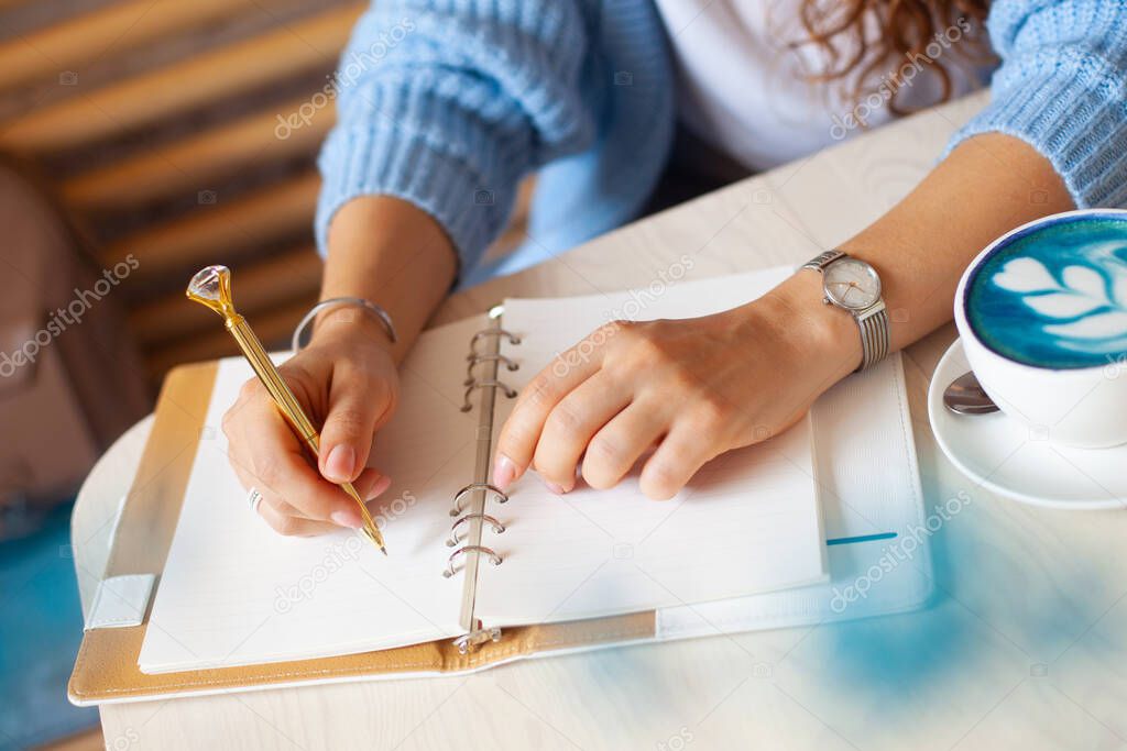Well groomed woman hand holding gold pen and writing notes with gold pen in notebook while drinking blue latte beside window. Freelance journalist working at home. Planning future concept. Copy space