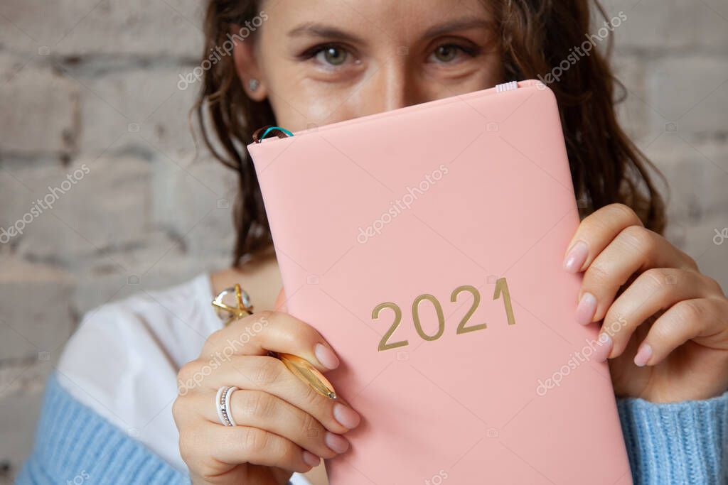 Woman with wavy hair is closing her face with a pink coloured planner 2021 near beige brick wall. New year planning concept with future ideas and goals. Copy space
