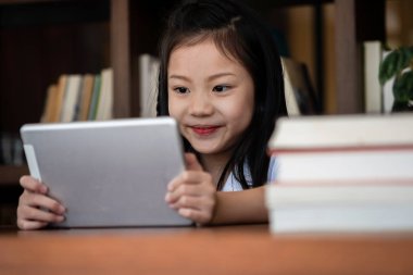 Şirin kız gülümsüyor ve kütüphanede tablet akıllı telefon oynuyor. Çocuk konsepti, eğitim konsepti.