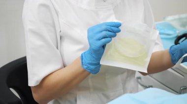 Mavi koruyucu eldivenli bir diş hekiminin, içinde tek kullanımlık ağız retraktörü bulunan steril bir paketi açması. Modern bir diş sağlığı kliniği.