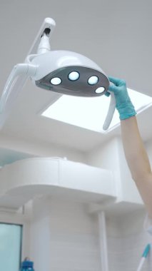 Dişçide genç bir kadın. Diş hekimi modern bir diş kliniğindeki hasta randevusu için diş lambasını ayarlar. Dikey video.
