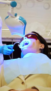 Modern bir diş kliniğinde morötesi diş beyazlatma prosedürü. Doktor hastaların dişlerini beyazlatır. Kozmetik dişçiliği. Dikey video.