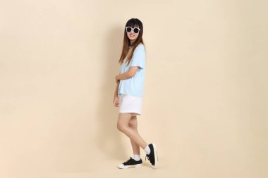 Asyalı bir kadın mavi polo gömlek, beyaz şort, güneş gözlüğü takıyor, kolları çapraz duran ve güvenle bej bir arka plana gülümseyen. Üniversite öğrencisi, Gen Z, Summer