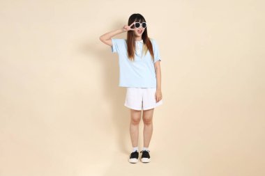 Asyalı bir kadın mavi polo gömlek, beyaz şort, güneş gözlüğü, barış işareti, V işareti, iki parmağı bej arka planda. Üniversite öğrencisi, Gen Z, Summer