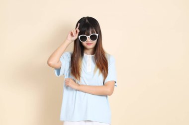 Asyalı bir kadın mavi polo gömlek, beyaz şort, güneş gözlüğü giyiyor, bej arka planda düşünüyor. Üniversite öğrencisi, Gen Z, Summer