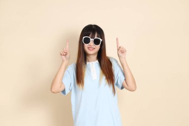 Asyalı bir kadın mavi polo gömlek, beyaz şort, güneş gözlüğü takıyor, bej bir arka planı işaret ediyor. Üniversite öğrencisi, Gen Z, Summer