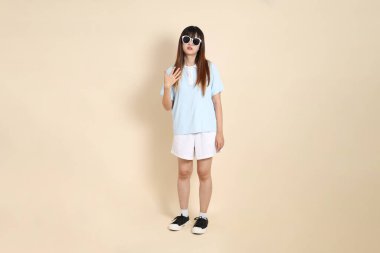 Mavi polo tişörtlü, beyaz şortlu, güneş gözlüklü, bej arka planda sıcak hisseden Asyalı bir kadın. Üniversite öğrencisi, Gen Z, Summer