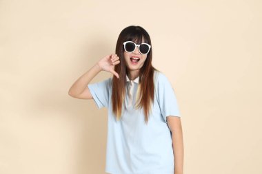 Asyalı kadın mavi polo gömlek, beyaz şort, güneş gözlüğü, bej arka planda baş parmak izi. Üniversite öğrencisi, Gen Z, Summer