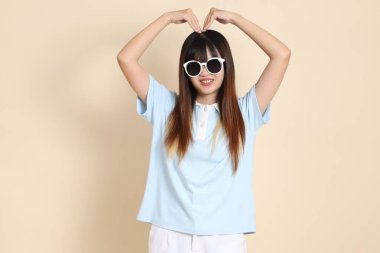 Asyalı bir kadın mavi polo gömlek, beyaz şort, güneş gözlüğü, kalp şeklinde mini kalp, bej arka planda aşk. Üniversite öğrencisi, Gen Z, Summer