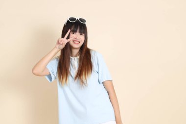 Asyalı bir kadın mavi polo gömlek, beyaz şort, güneş gözlüğü, barış işareti, V işareti, iki parmağı bej arka planda. Üniversite öğrencisi, Gen Z, Summer