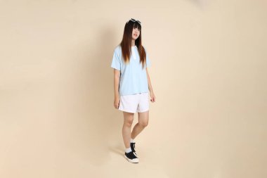 Mavi polo tişörtlü, beyaz şortlu, güneş gözlüklü, bej arka planda yürüyen Asyalı bir kadın. Üniversite öğrencisi, Gen Z, Summer