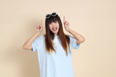 Asyalı bir kadın mavi polo gömlek, beyaz şort, güneş gözlüğü takıyor, bej bir arka planı işaret ediyor. Üniversite öğrencisi, Gen Z, Summer