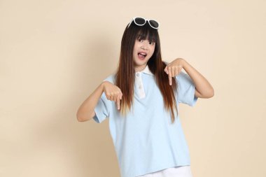 Asyalı bir kadın mavi polo gömlek, beyaz şort, güneş gözlüğü takıyor, bej bir arka planı işaret ediyor. Üniversite öğrencisi, Gen Z, Summer