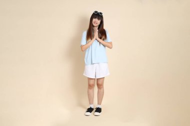 Mavi polo tişörtlü, beyaz şortlu, güneş gözlüklü, bej arkaplanlı Asyalı bir kadın. Üniversite öğrencisi, Gen Z, Summer