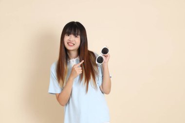 Asyalı bir kadın mavi polo gömlek, beyaz şort, güneş gözlüğü takıyor, bej bir arka planı işaret ediyor. Üniversite öğrencisi, Gen Z, Summer