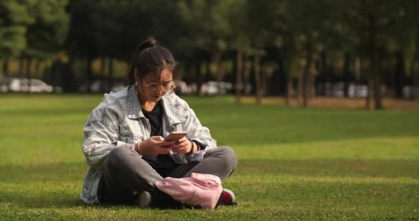 Asiática chica universitaria sentado en el césped usando el teléfono móvil en el campus — Vídeos de Stock