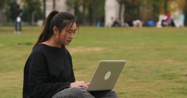 Asijská studentka, která sedí na trávníku a používá přenosný počítač v areálu — Stock video