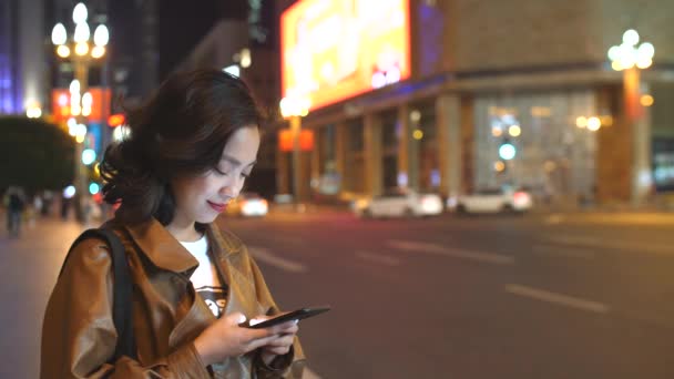 漂亮的年轻亚洲妇女使用手机在城市街道在晚上，4k — 图库视频影像