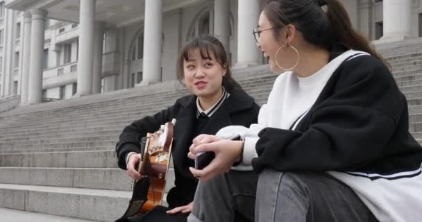 Две молодые девушки поют вместе счастливо на ступеньках с гитарой — стоковое видео