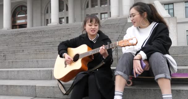 Две китайские девушки учатся играть на гитаре и петь в кампусе — стоковое видео