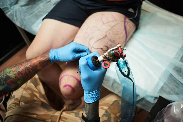 dark-haired tattooist holding tattoo machine and making tattoo to client in workshop