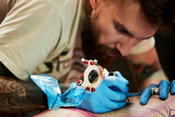 concentrated man tattoo master making tattoo on leg of client by tattoo machine, close-up
