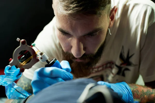 Young dark-haired man tattoo master making tattoo to client by tattoo machine, close-up