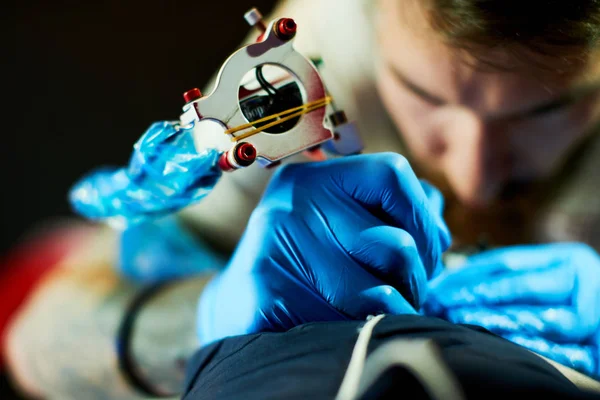 Young dark-haired man tattoo master making tattoo to client by tattoo machine, close-up