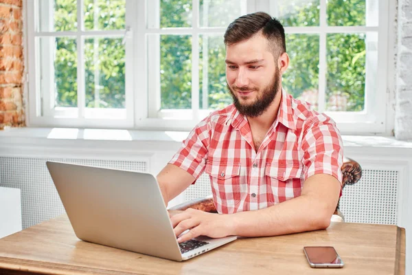 handsome bearded man and working on laptop while sitting at table in room near windows