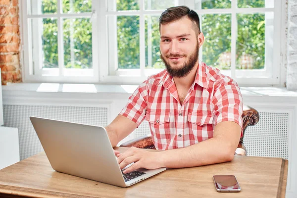 handsome bearded man looking at camera and working on laptop while sitting at table in room near windows