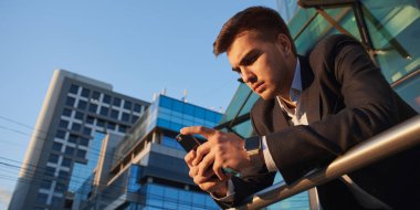 arka planda bina ile korkuluk üzerine eğilerek süre smartphone kullanarak takım elbiseli yakışıklı işadamı