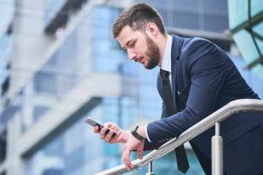 arka planda bina ile korkuluk üzerine eğilerek süre smartphone kullanarak takım elbiseli yakışıklı zarif işadamı