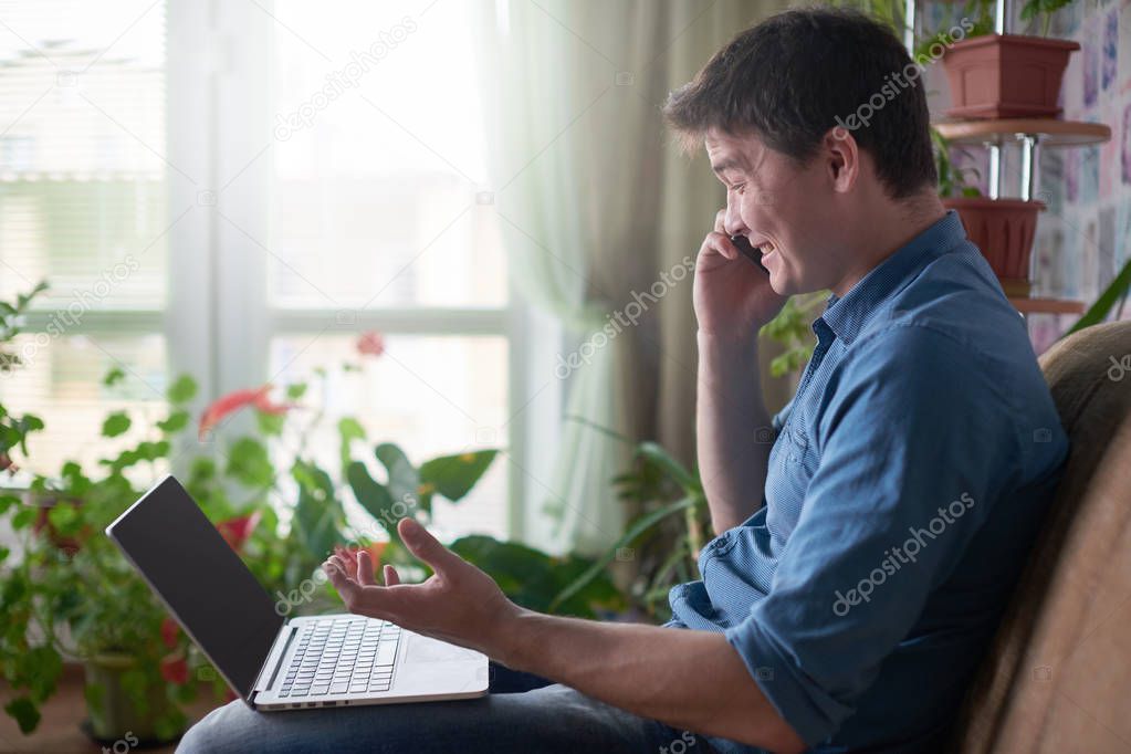 man talking on smartphone while sitting on sofa with laptop at home 