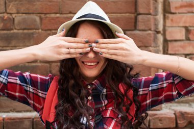 elleriyle gözlerini kapatma ve kırmızı tuğla duvar arka plan üzerinde gülümseyen şapkalı kadın 