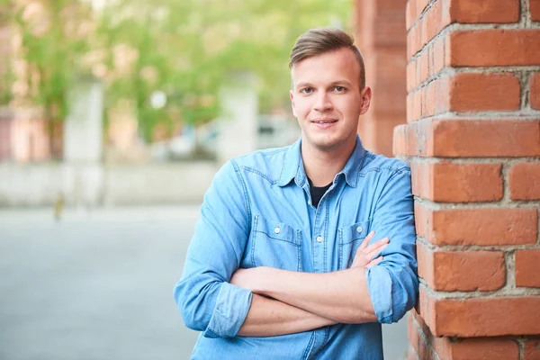 student in denim shirt with crossed arms looking at camera and leaning on orange brick building