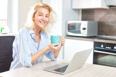 kahve fincanı tutarak ve mutfak, kahvaltı kavramı içinde laptop ile masada oturan sevimli kadın 