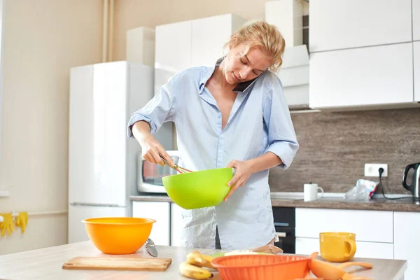 woman holding whisk and mixing ingredients in bowl while talking on phone at kitchen, close-up