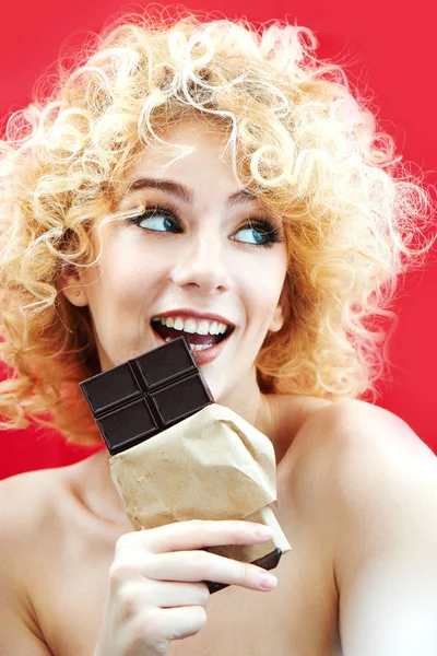 beautiful woman with curly hair biting chocolate bar and smiling on red background