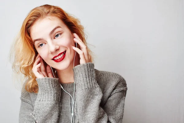 セーターは 灰色の背景の上にヘッドフォンで音楽を聴く若い赤髪の女性の肖像画 — ストック写真