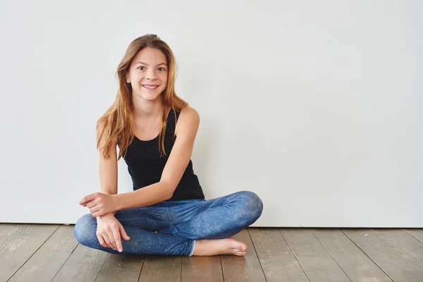 girl in black tank top and jeans sitting barefoot on wooden floor against white background