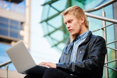 deri ceket ve basamaklarında oturan ve arka plan üzerinde şehir binaları ile dizüstü bilgisayarda yazarak gömlek sakallı sarışın adam 
