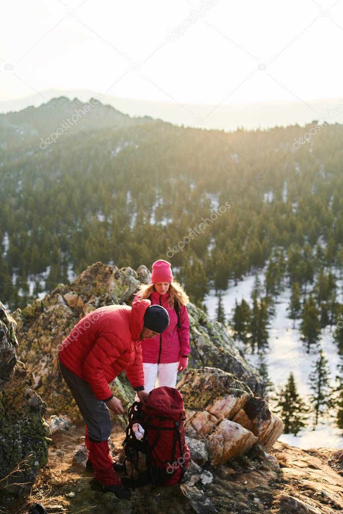 man straightening backpack while standing with woman on top of sunlit mountain 