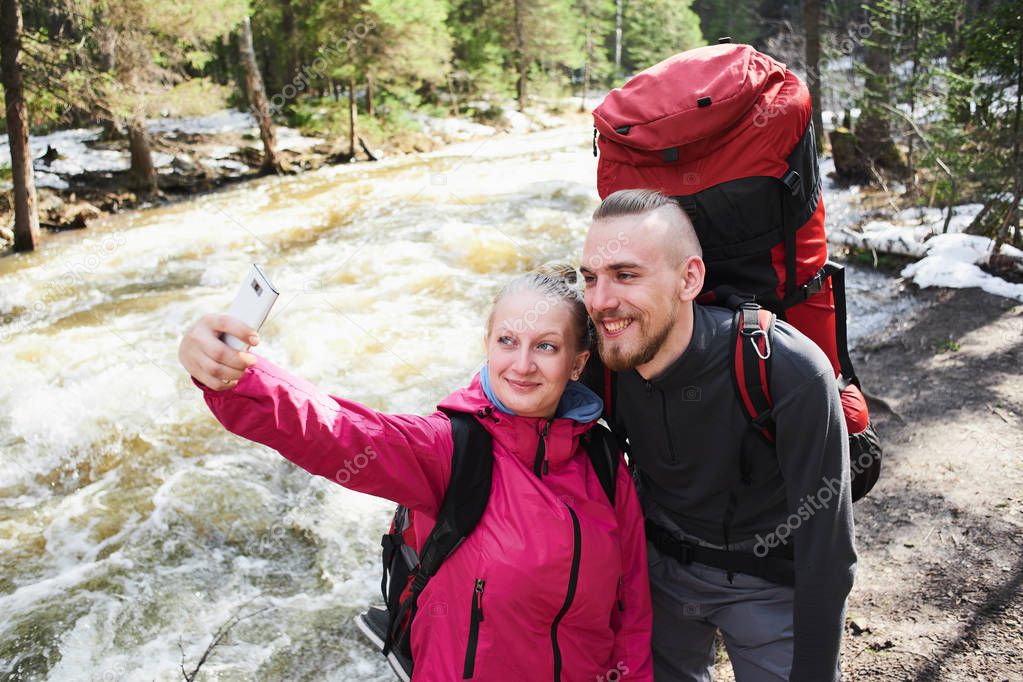 tourist couple in love taking selfies with smartphone near flowing river in forest 