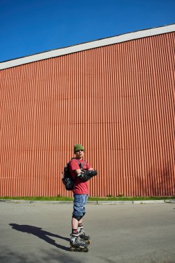 Tekerlekli paten ayakta kırmızı duvarının yakınında çapraz kolları olan kendine güvenen adam 