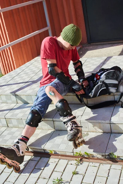 man in roller skates with abrasions on his knee sitting on stairs and searching in backpack tools for first aid