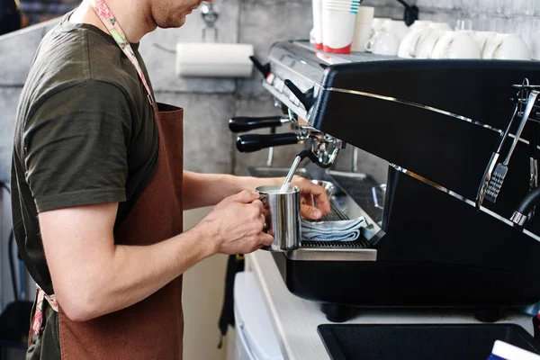 Barista steaming milk in pitcher with coffee machine while preparing to make latte in cafe