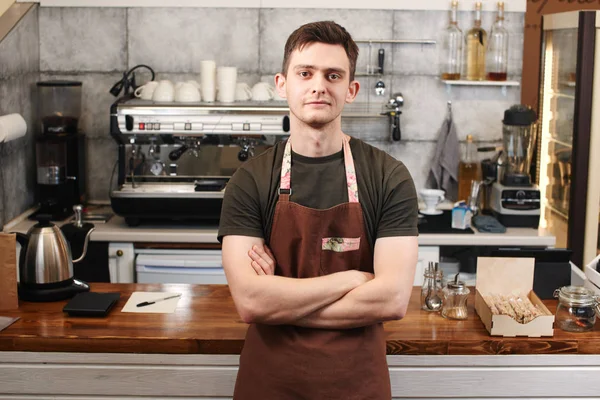 man barista with crossed arms leaning on wooden bar surface at workplace in coffee shop