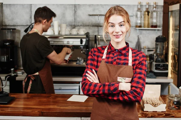 female barista with crossed arms looking at camera while standing in front of bar with colleague near coffee machine on background