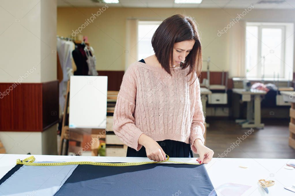 concentrated seamstress putting textile on table and measuring with tape, creation of clothes in atelier concept