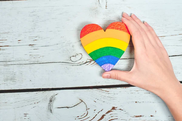 female hand holding heart painted in rainbow colors on wooden background, LGBT concept
